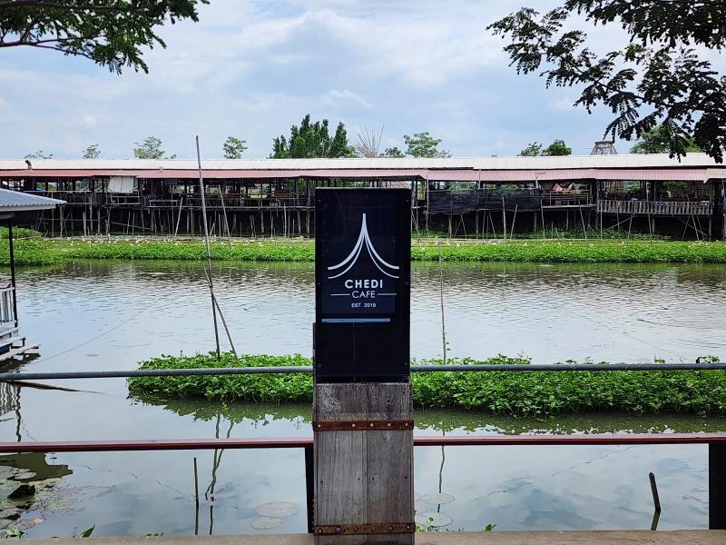The black Chedi Cafe sign on a wooden post at the canal rail, with floating weed and a covered stilt market on the far bank.