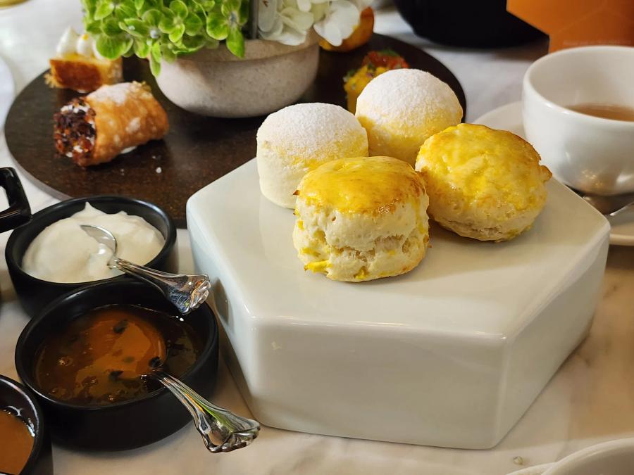 Four small scones on a white hexagonal stand, two glazed and two dusted with icing sugar, beside bowls of cream and passion fruit jam.