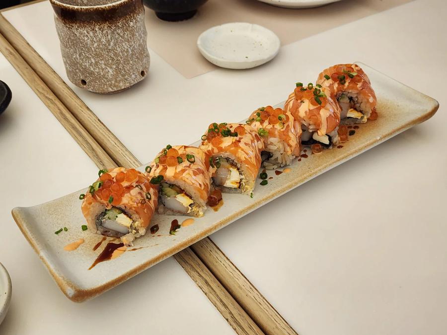 Six pieces of salmon-topped sushi roll on a long speckled plate, each topped with salmon roe and spring onion, with drizzles of spicy and dark sauce.