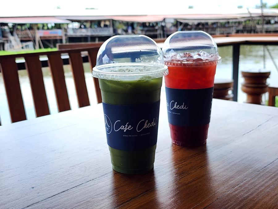 Two iced drinks in plastic cups with blue Cafe Chedi sleeves on a wooden table, a green matcha in front and a red plum tea behind, with a canal and a stilt market out of focus beyond the rail.