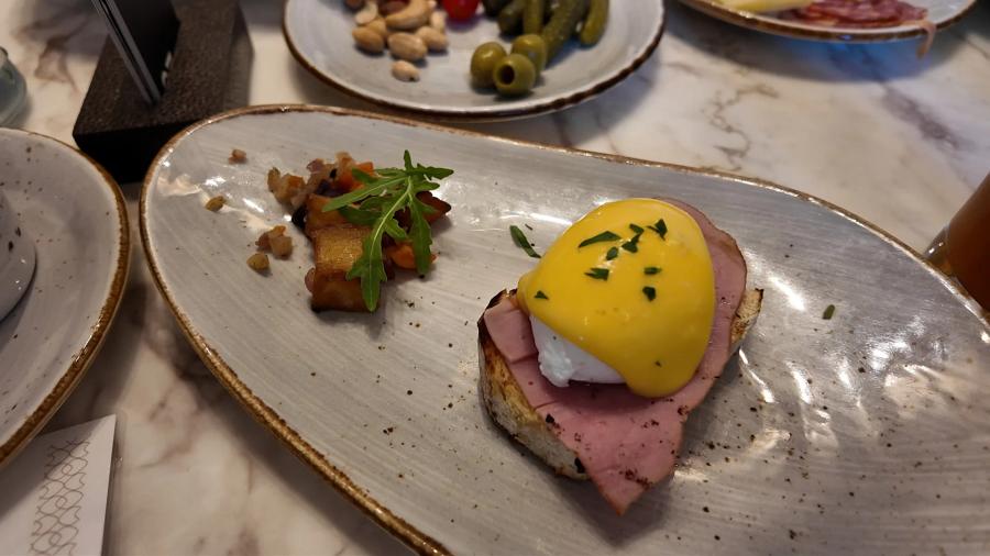 Eggs Benedict with ham and hollandaise on toasted bread beside grilled tomato and rocket on a grey plate at Bella Sera.