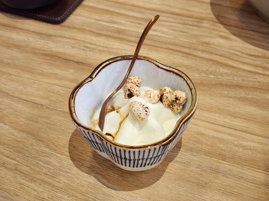 A small ceramic bowl of white milk pudding with dark syrup and caramel popcorn on top, with a copper spoon resting in it.