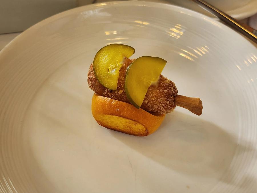 A small crumbed chicken drumstick topped with two slices of pickle, resting on a split brioche roll on a white plate.