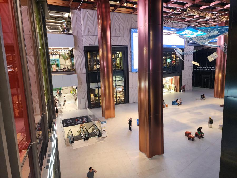 The atrium of The Storeys at One Bangkok seen from above, with copper columns, an escalator signed Food Parade and shoppers crossing the stone floor.