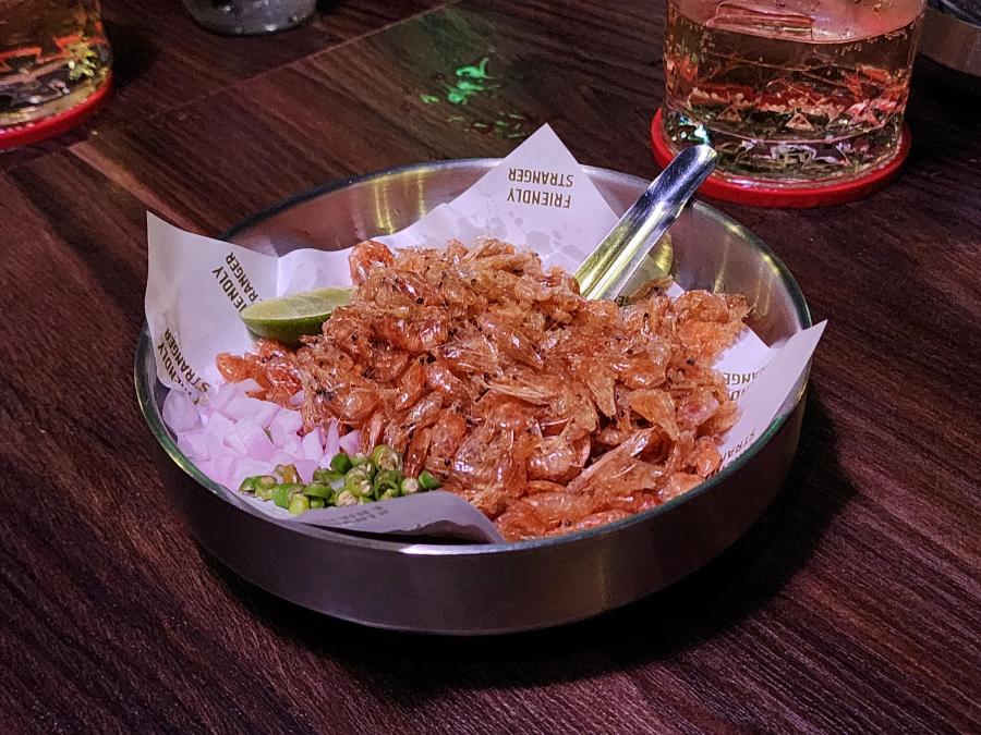 A steel tray of small crispy fried shrimp with diced shallots, sliced green chillies and a lime wedge, with two glasses on red coasters behind.