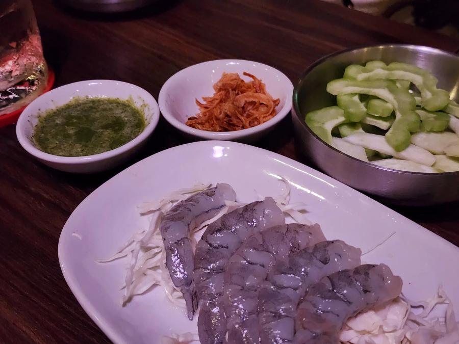 Raw butterflied prawns on shredded cabbage, with a bowl of green dipping sauce, a bowl of crispy fried topping and a steel tray of sliced bitter melon.