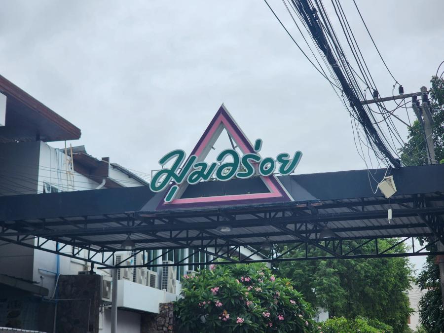 The Moom Aroi name in green Thai script on a pink and purple triangle above a steel-framed gate, under a grey sky.