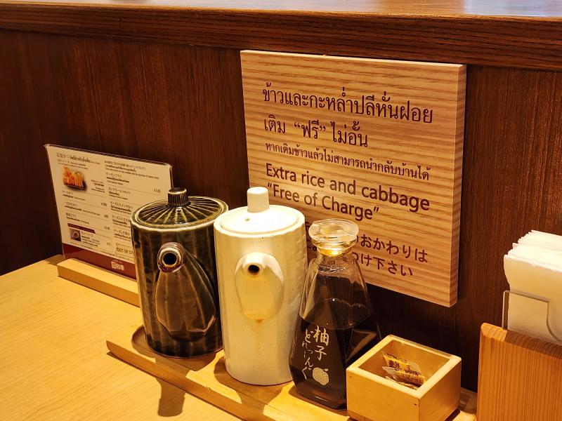 Two ceramic sauce pots, a bottle of yuzu dressing and a box of sesame sachets on a wooden tray under the free refill sign at Tonkatsu Wako.