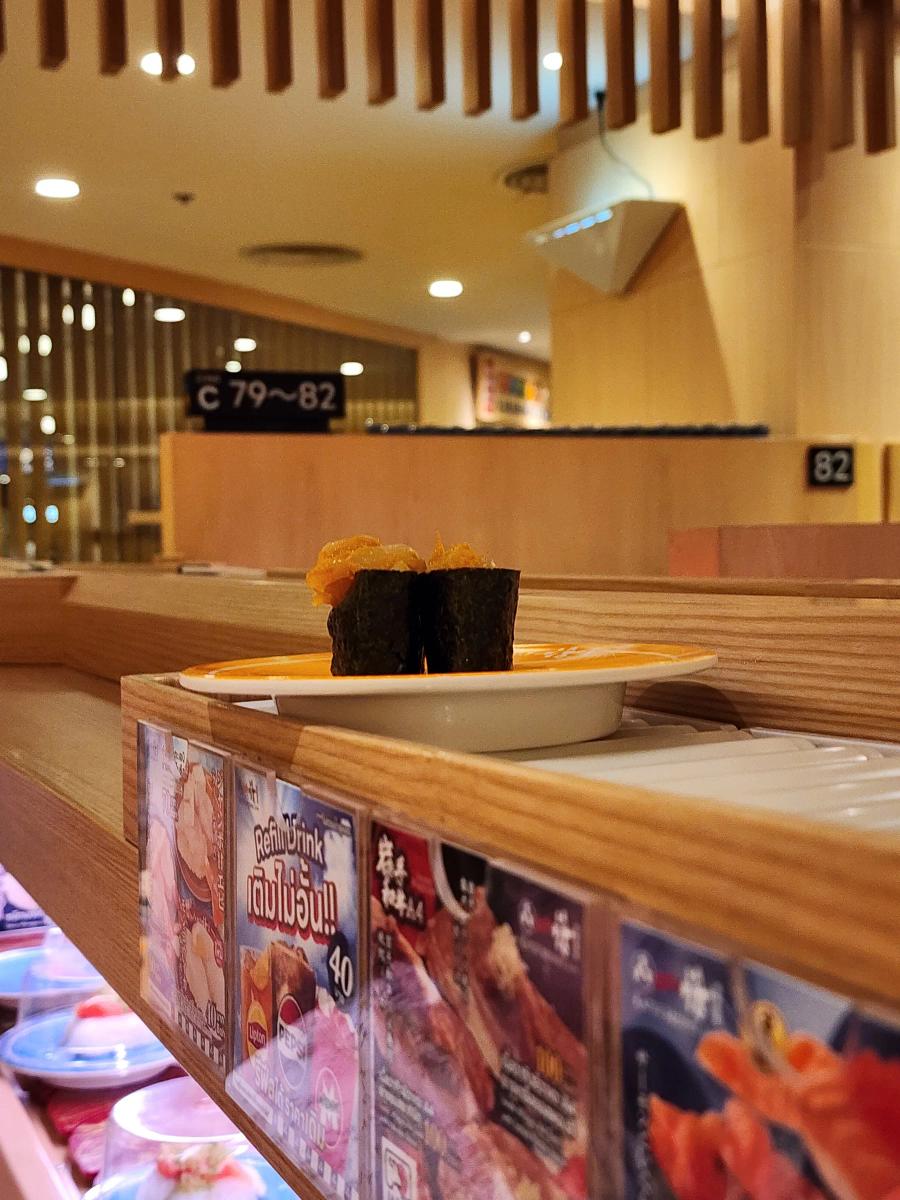 Two gunkan rolls with an orange topping on an orange plate moving along the belt, with counter seats numbered C79 to 82 behind.