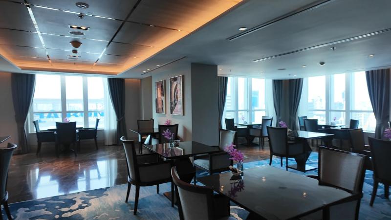 The dining room of the Club Millésime lounge with dark tables, orchids, a blue patterned carpet and windows on two sides over Bangkok.