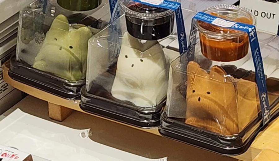 Three cat-shaped puddings in clear takeaway domes, one green, one white and one caramel brown, each with a pot of sauce on top.