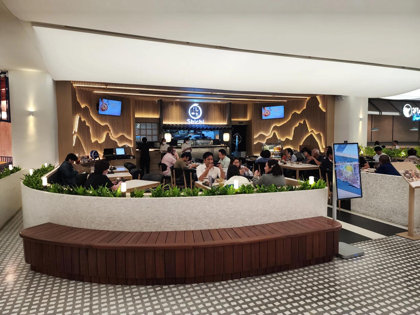 The front of Shichi Japanese Restaurant at Central Chidlom, with its round sign over the open kitchen, backlit mountain panels on a slatted wood wall, a low planter wall and a wooden bench outside.