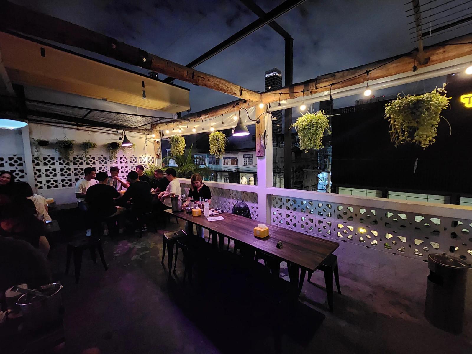 The rooftop terrace at Friendly Stranger at night, with a long timber table, white breeze block walls, hanging plants and a group drinking at the far end.