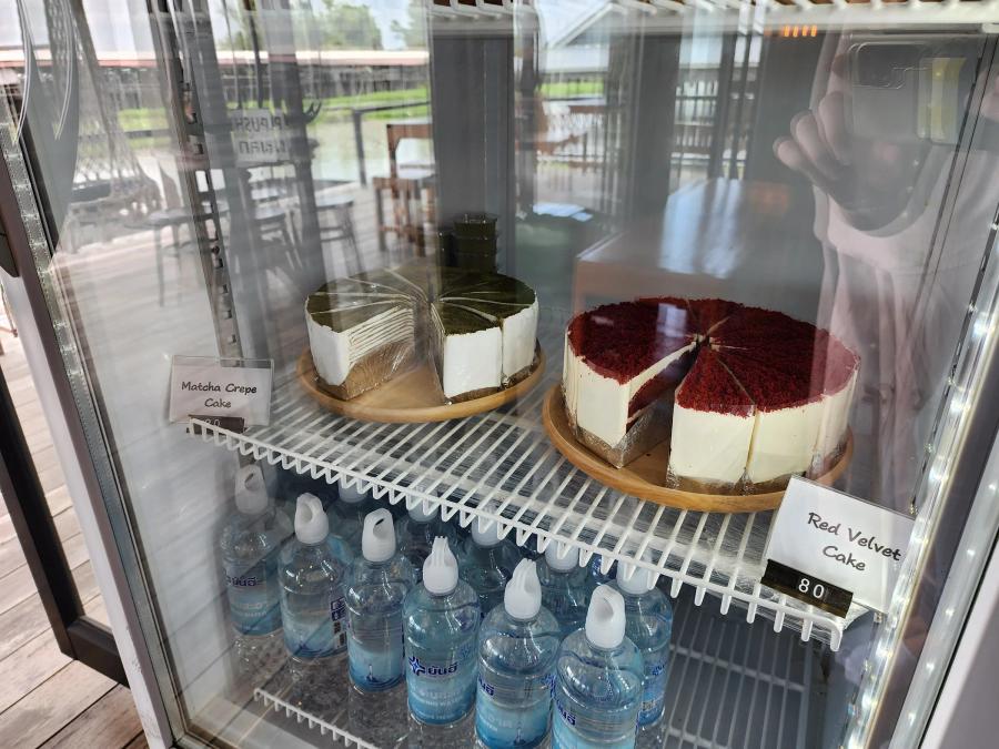 A glass fridge at Cafe Chedi holding a sliced matcha crepe cake and a sliced red velvet cake on wooden boards above rows of bottled water.