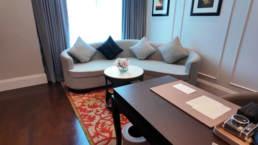 The living room of a Junior Suite at Sofitel Bangkok Sukhumvit with a curved cream sofa, cushions, a round coffee table and a red patterned rug on dark wood flooring.