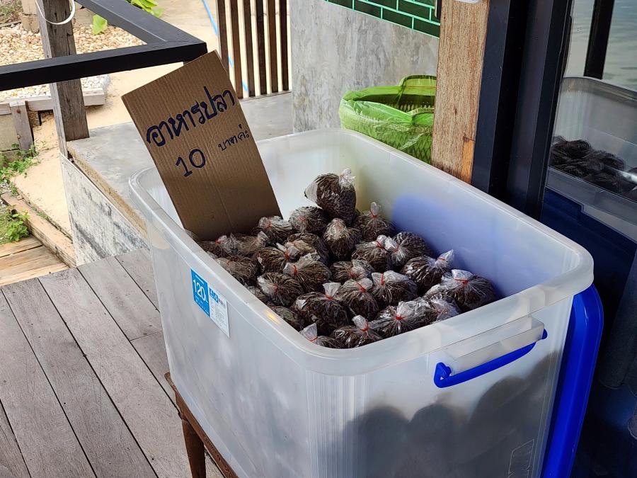 A clear plastic tub of small bags of fish pellets on the deck at Cafe Chedi, with a cardboard sign reading ten baht in Thai.