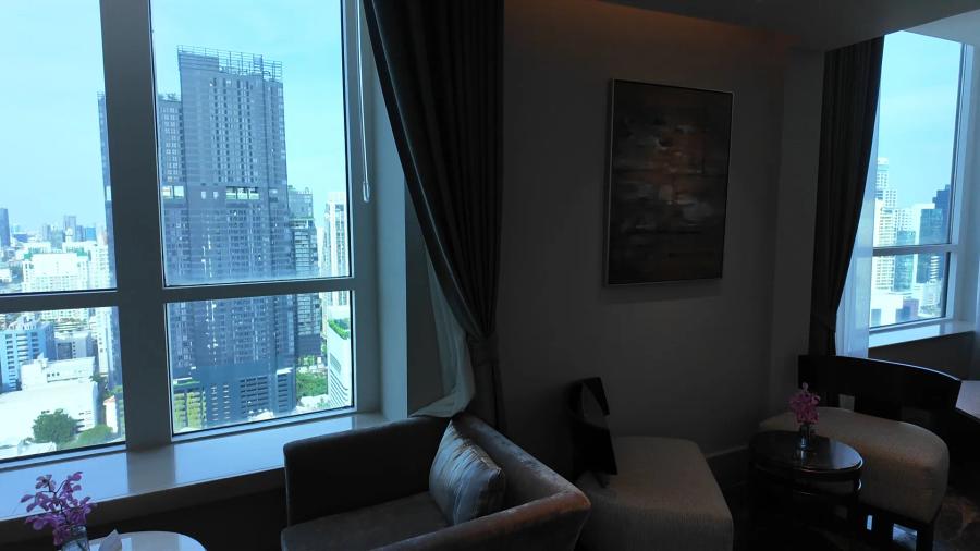 A grey sofa and armchair beside tall windows in the Club Millésime lounge, with an orchid on a side table and Bangkok towers outside.
