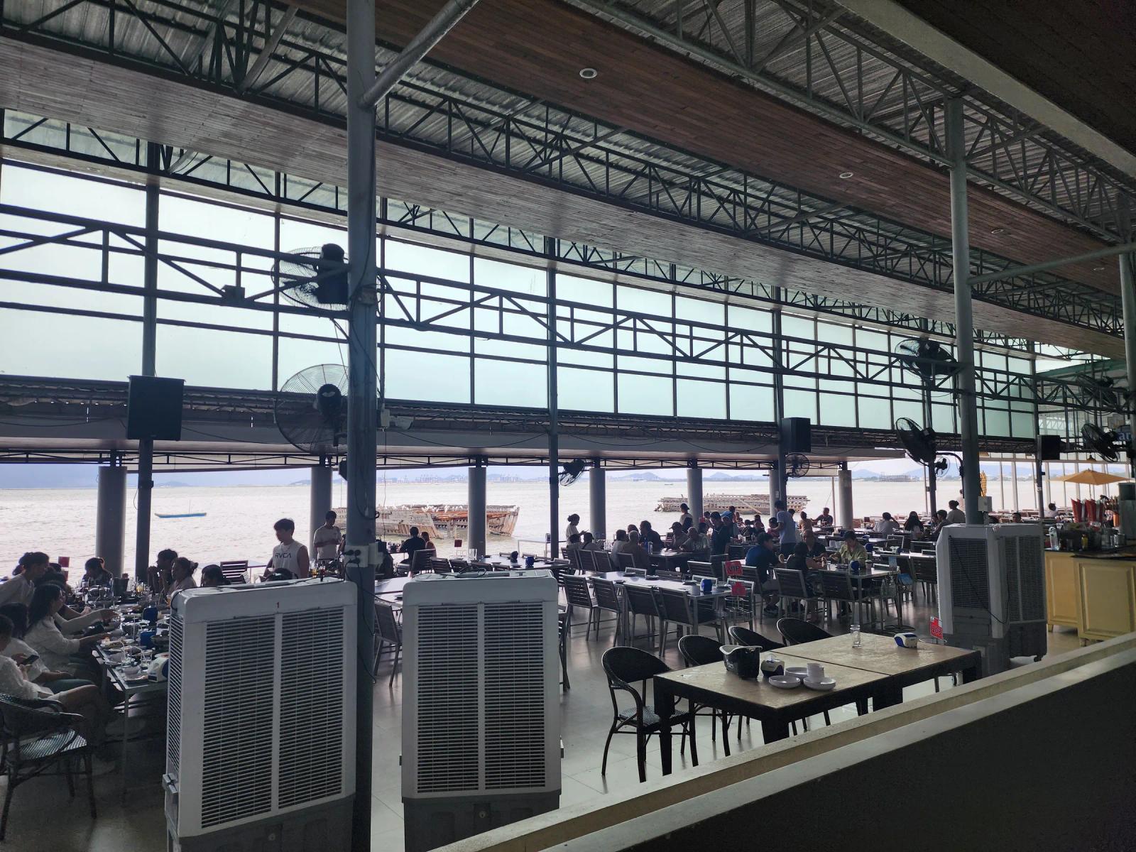 The open-sided dining hall at Moom Aroi in Naklua, Pattaya, with rows of tables under a steel roof, cooling fans and the sea beyond.