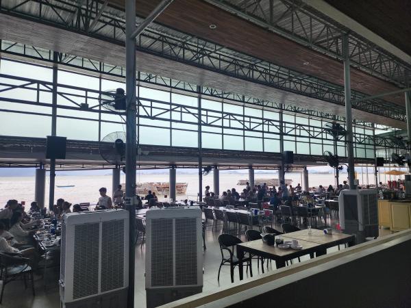 The open-sided dining hall at Moom Aroi in Naklua, Pattaya, with rows of tables under a steel roof, cooling fans and the sea beyond.