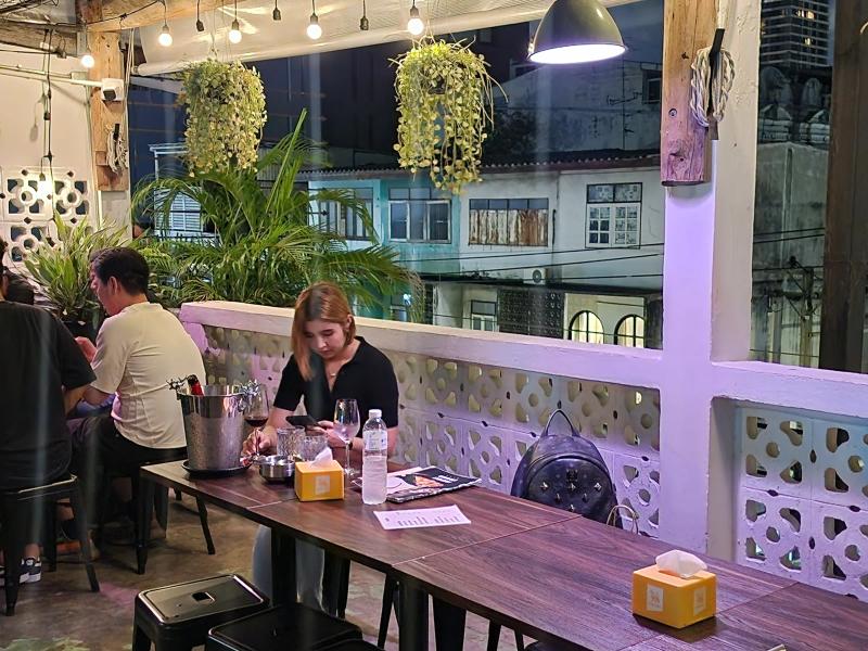A corner of a rooftop terrace with white breeze block walls, hanging plants under a string of bulbs and customers at a long timber table.