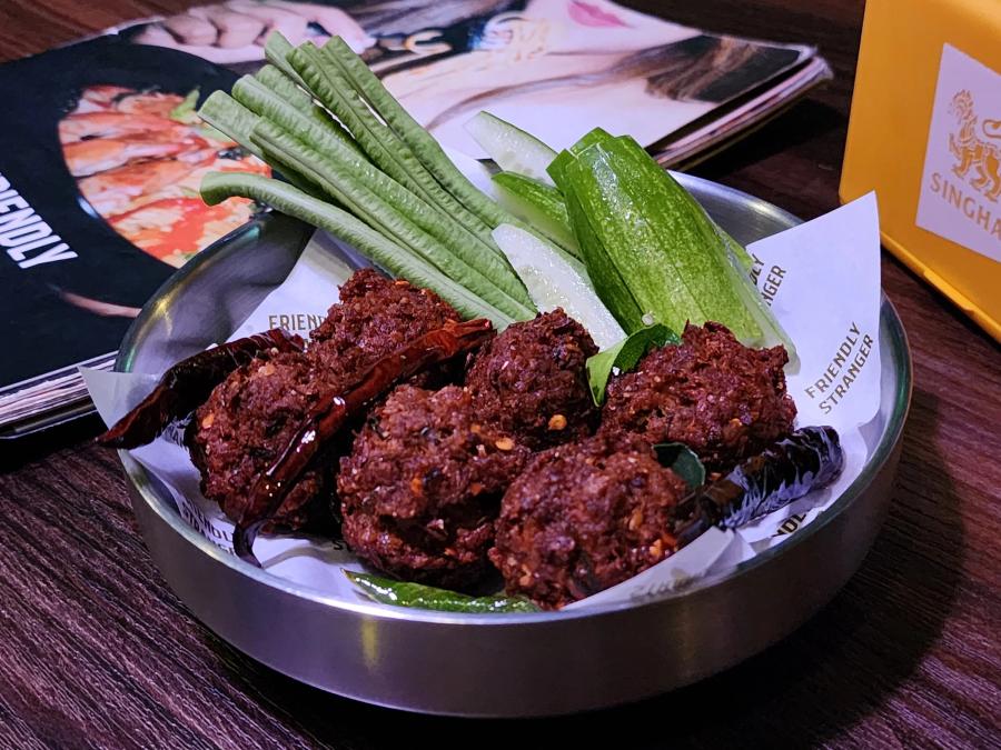 Dark fried pork balls with whole dried chillies in a steel tray lined with Friendly Stranger paper, beside raw long beans and cucumber.