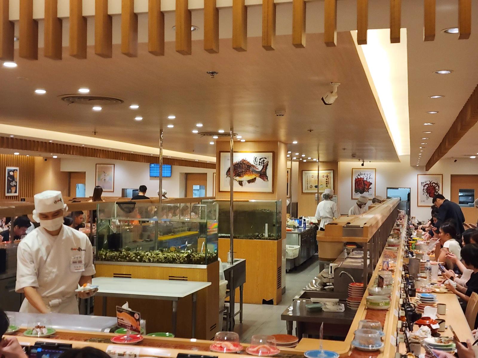 The long sushi counters at Katsu Midori CentralWorld, with chefs in white caps working beside glass tanks, framed fish prints on the walls and diners along the belt.
