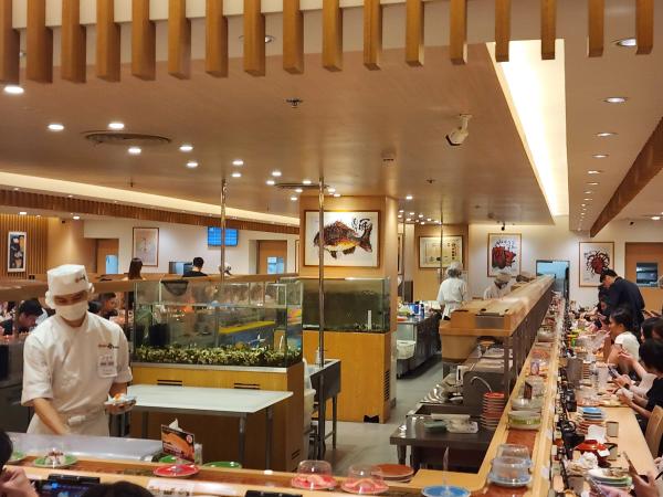 The long sushi counters at Katsu Midori CentralWorld, with chefs in white caps working beside glass tanks, framed fish prints on the walls and diners along the belt.