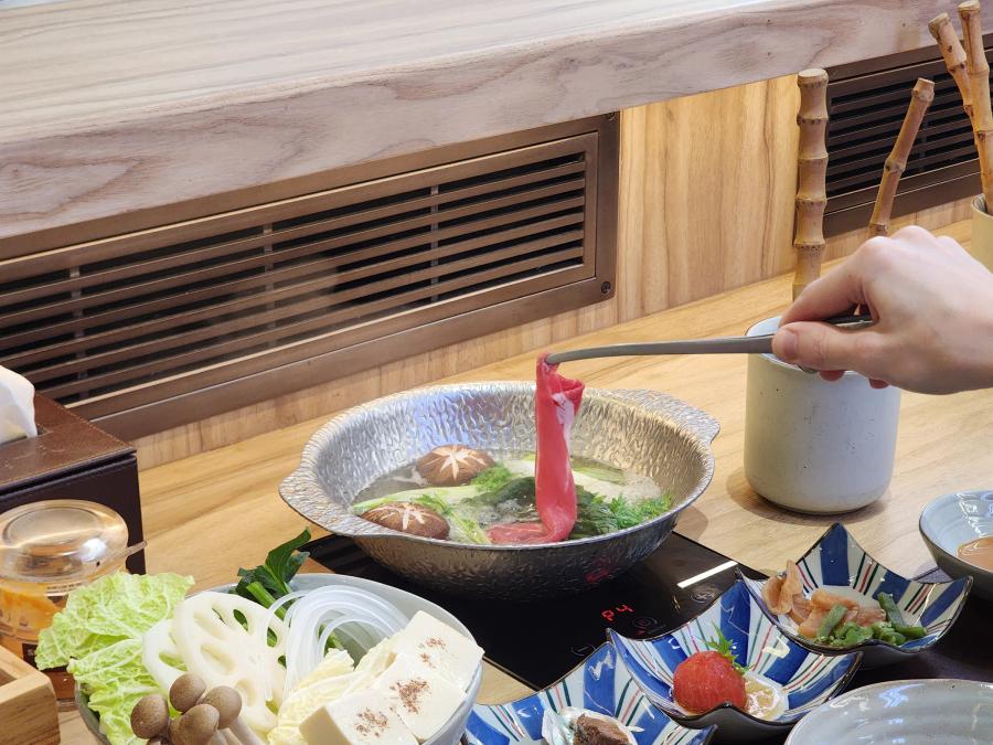 A hand holding a slice of red Charolais beef in tongs over a hammered metal pot of broth, with a bowl of vegetables and three starter dishes on the counter below.
