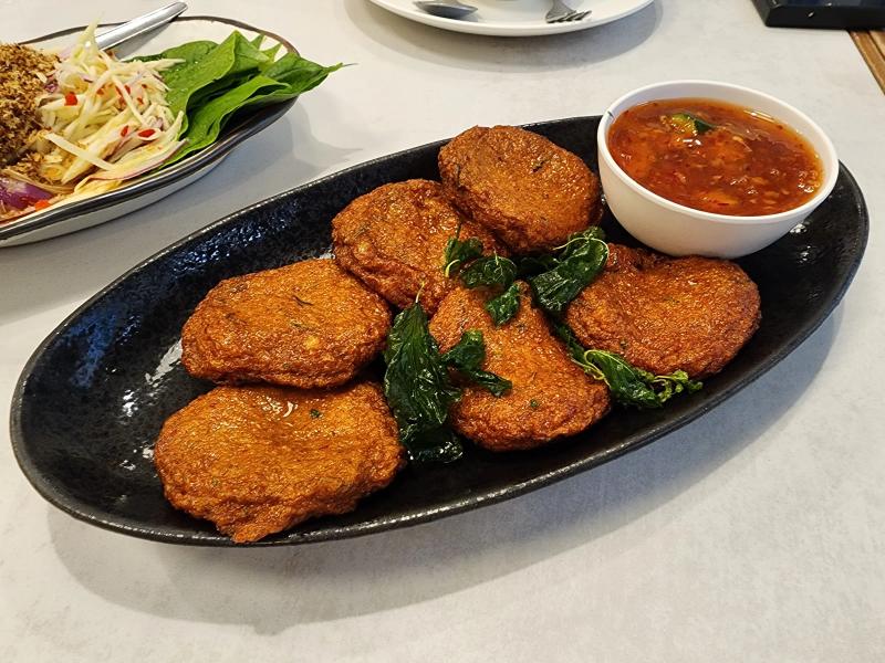 Six fried fish cakes with fried basil leaves and a bowl of sweet chilli dip on a black oval plate, with the mango salad on the table behind.