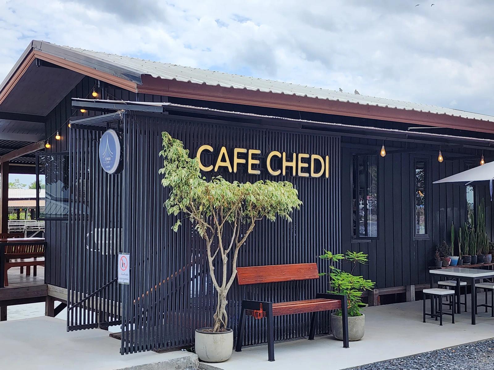 The black timber front of Cafe Chedi with gold lettering, a wooden bench, a potted tree and stools under a white umbrella.
