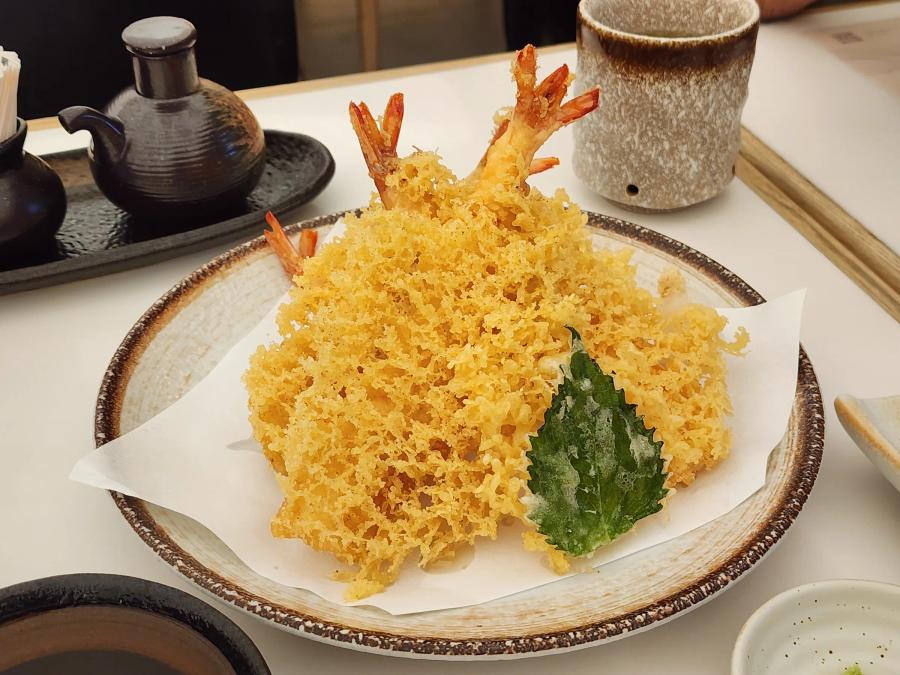 Three prawn tempura in a lacy golden batter with a tempura shiso leaf on a round speckled plate lined with paper.