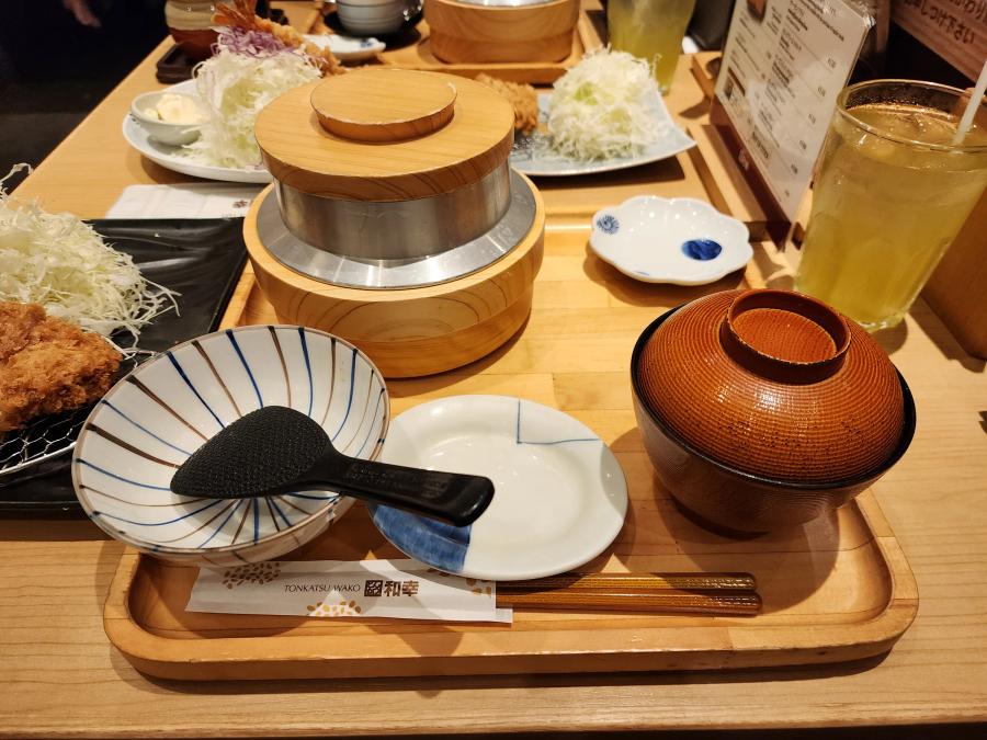 A wooden tray at Tonkatsu Wako with a lidded wooden rice pot, a striped mortar holding a black pestle, a small dish, a lidded red miso soup bowl and chopsticks in a paper sleeve.