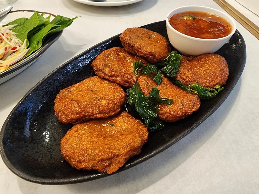 Six thick fried fish cakes on a black oval plate with fried basil leaves and a bowl of sweet chilli dip with cucumber.