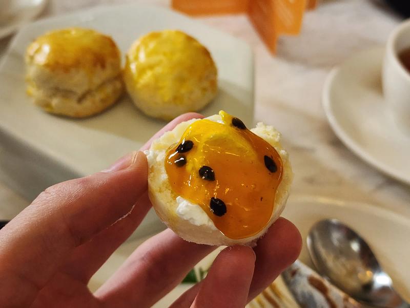 A hand holding half a small scone spread with cream and orange passion fruit jam with black seeds, two glazed scones behind.