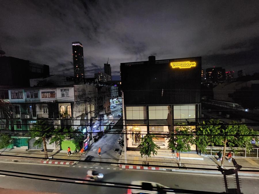 A night view from a rooftop over a Bangkok road, with old shophouses, a side street, a black building with a yellow neon sign and a tower in the distance.