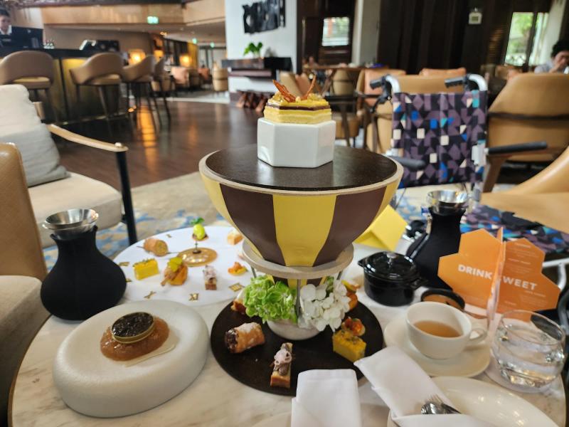 The hot air balloon stand with its top removed, a hexagonal honey mousse on a white stand above tiers of savouries and sweets, with the lounge of the Diplomat Bar behind.