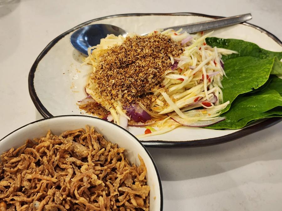 A green mango salad with shallot, chilli and a toasted crumb on a white plate beside a bowl of crisp fried shredded catfish.