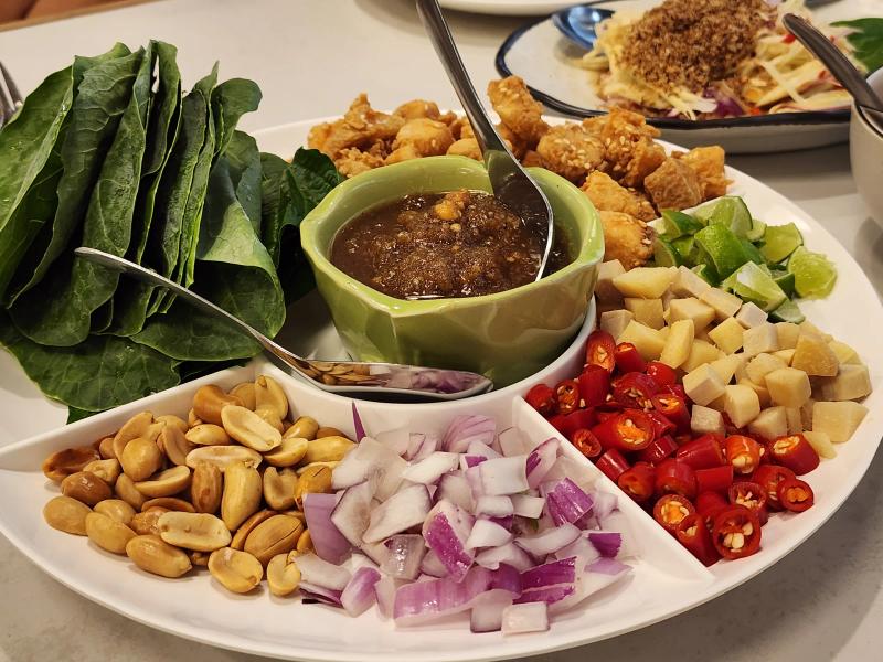 A close view of the leaf wrap platter: leaves, fried sea bass in sesame batter, lime, ginger, chilli, shallot and roasted peanuts around a green bowl of dark sauce.