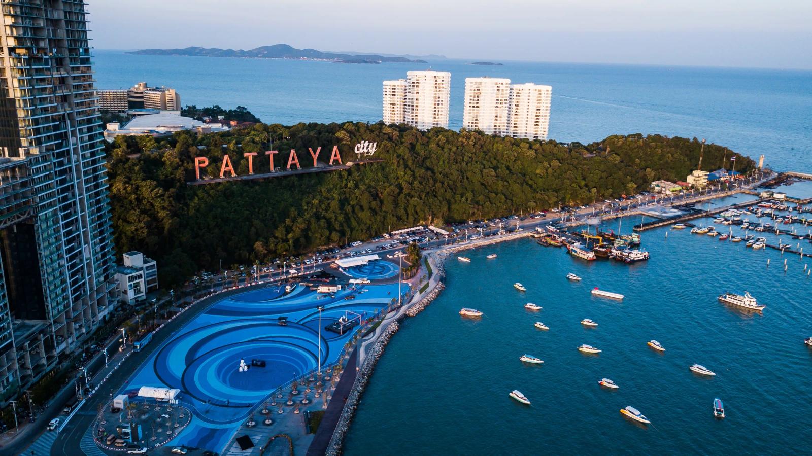 An aerial view of the Pattaya City sign on its wooded hill above the harbour, with boats moored in blue water and towers along the shore.