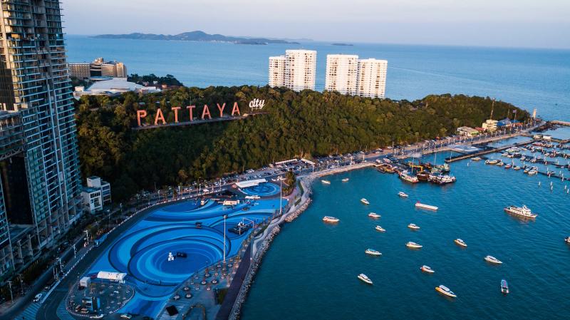 An aerial view of the Pattaya City sign on its wooded hill above the harbour, with boats moored in blue water and towers along the shore.