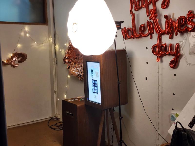 A wooden photo booth with a large round studio light, beside red foil balloons spelling Happy Valentines Day on a white wall.