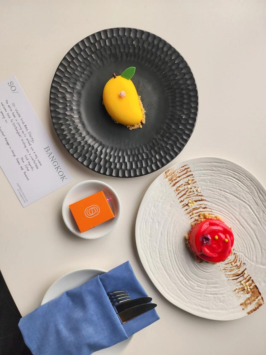 Mango-shaped dessert on a black plate and rose-shaped red dessert on a white plate beside a handwritten welcome card.