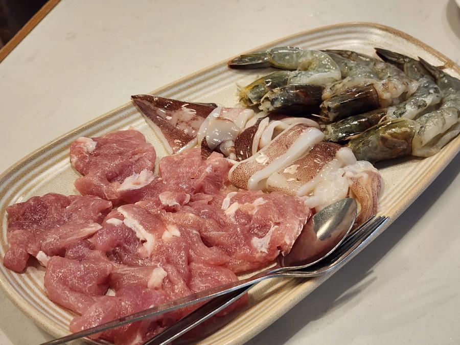 A platter of raw sliced pork, cuttlefish and whole prawns with a spoon and fork on a stone tabletop.