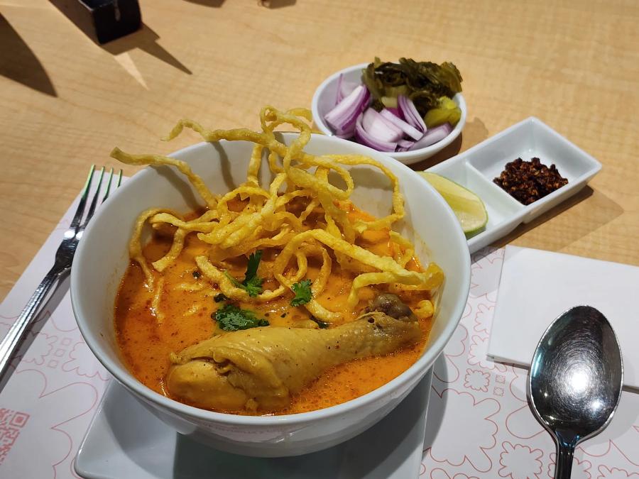 A bowl of khao soi with a chicken drumstick and crispy noodles, beside pickled mustard greens, shallots, lime and chilli.