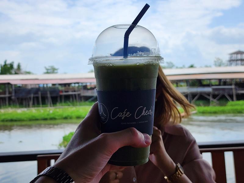 A hand holding up an iced matcha in a Cafe Chedi cup against the canal and the stilt market on the far bank.