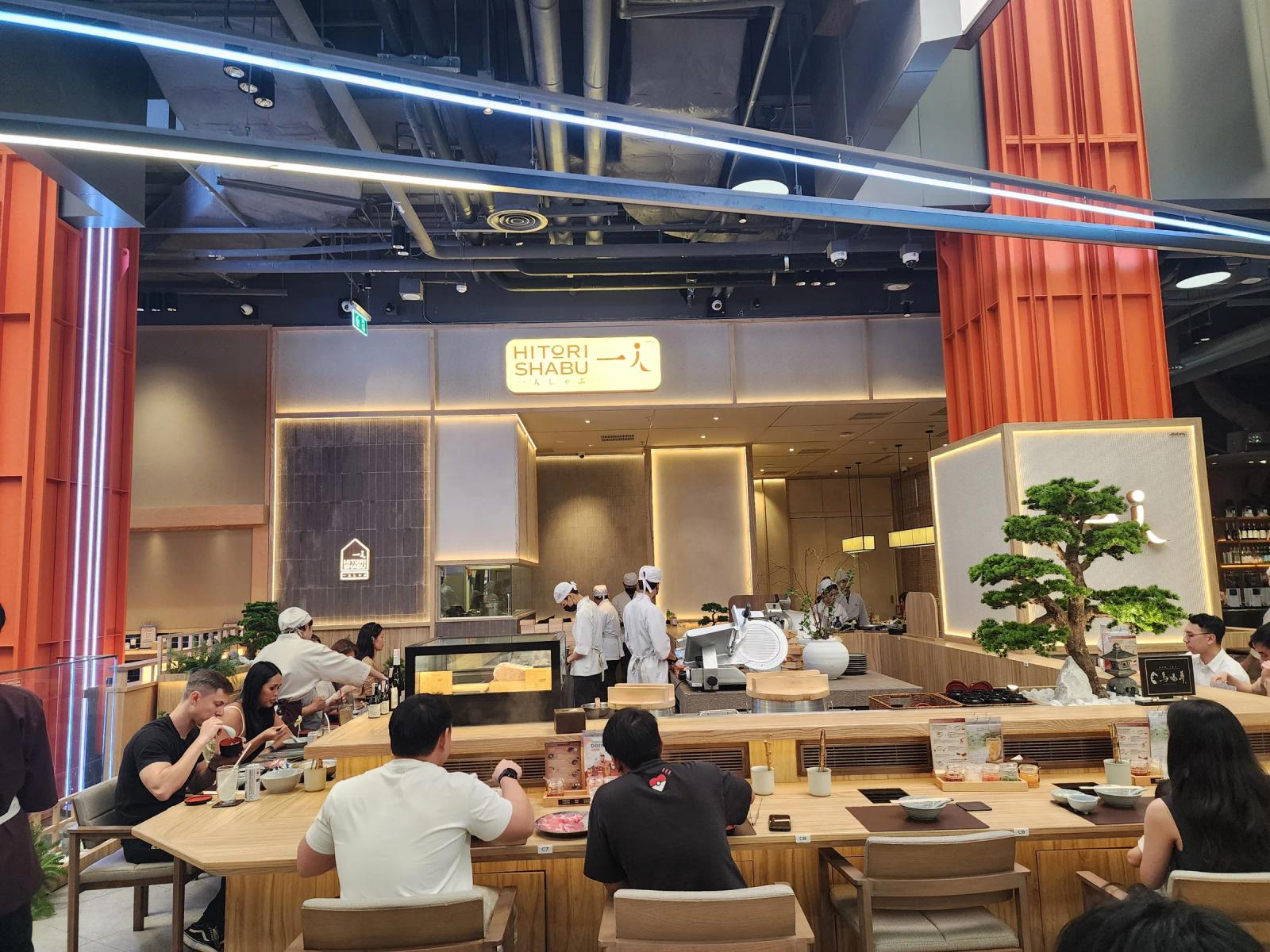 The front of Hitori Shabu One Bangkok, with its lit sign between two orange lattice pillars, chefs in white caps behind the open kitchen and diners along the wooden counter.