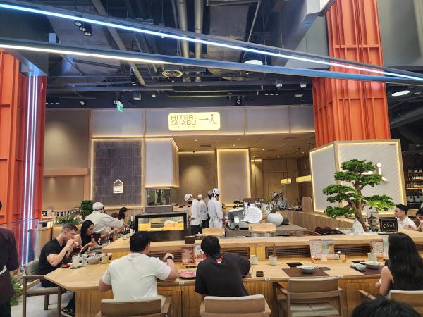The front of Hitori Shabu One Bangkok, with its lit sign between two orange lattice pillars, chefs in white caps behind the open kitchen and diners along the wooden counter.