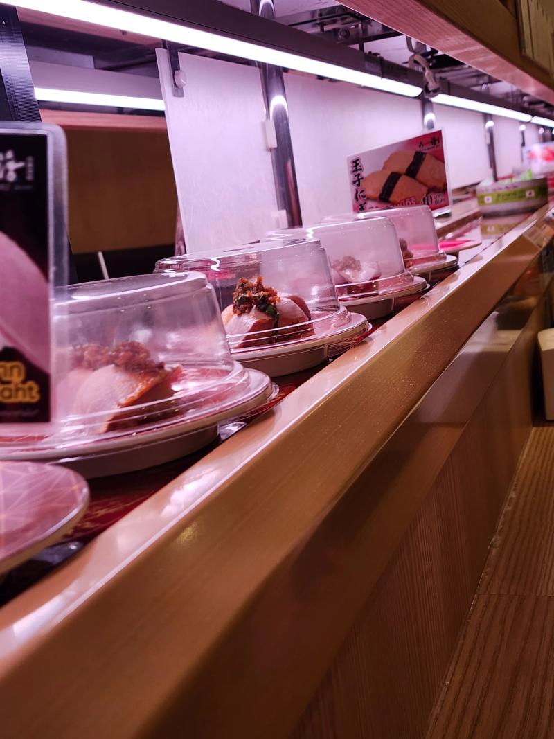 A row of domed sushi plates travelling along the belt under a pink-lit paper screen, with a card for tamago nigiri at 40 baht further along.