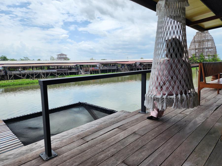 A person sitting in a white rope hammock chair on a wooden deck at Cafe Chedi, beside a black net platform over a canal, with a covered market on stilts on the far bank.