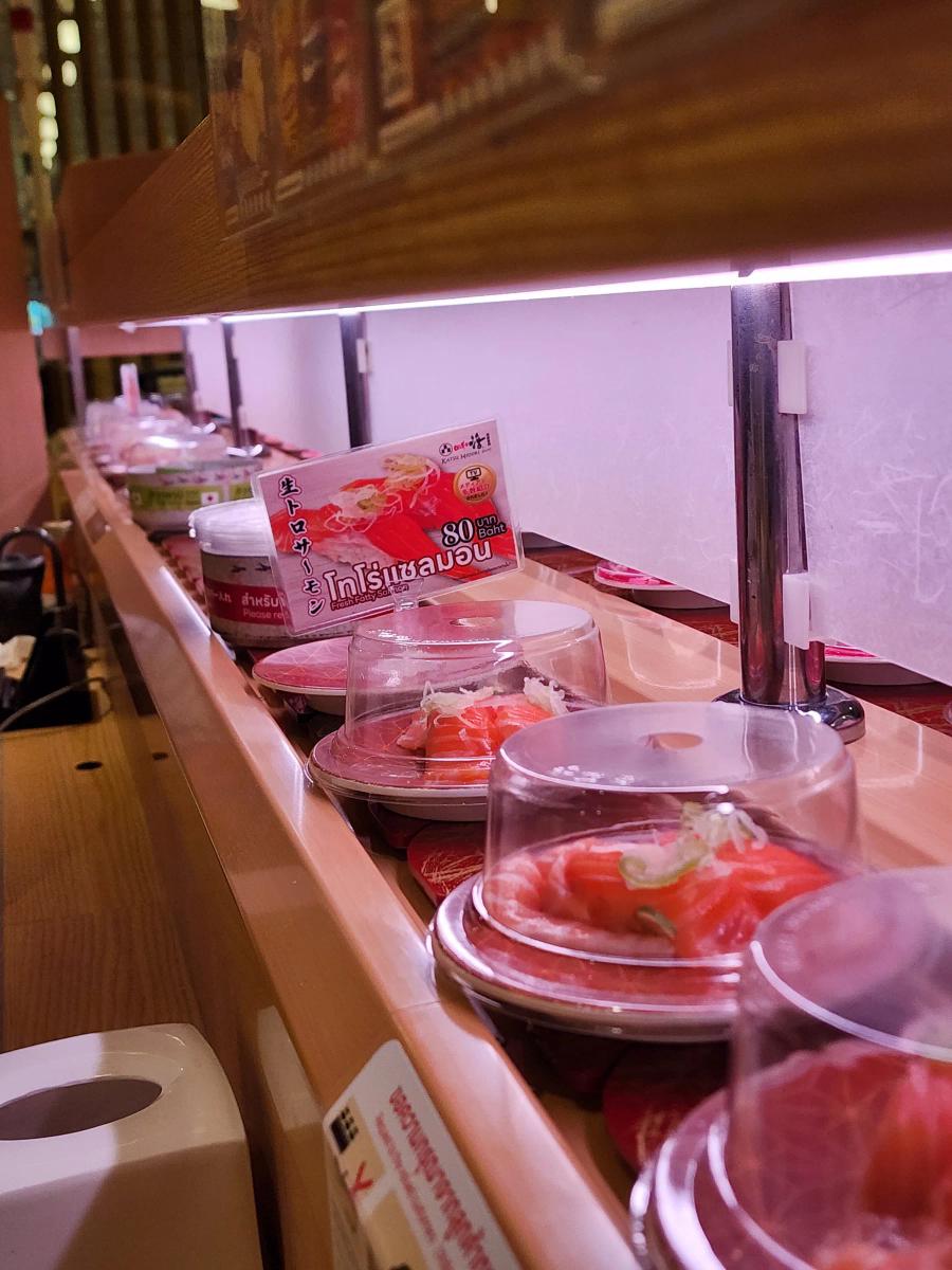 Domed plates of salmon nigiri moving along the conveyor belt at Katsu Midori, beside a card advertising fresh fatty salmon at 80 baht.